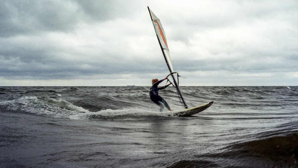 Windsurfer na wzburzonej, ciemnej wodzie pod zachmurzonym, szarym niebem. Żagiel jest biały z czerwonymi akcentami. W tle widoczny jest statek.