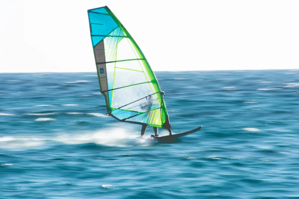 Windsurfer pędzący po turkusowej wodzie. Żagiel ma jaskrawe, neonowe kolory: niebieski i limonkowo-zielony, kontrastujące z jasnym niebem.